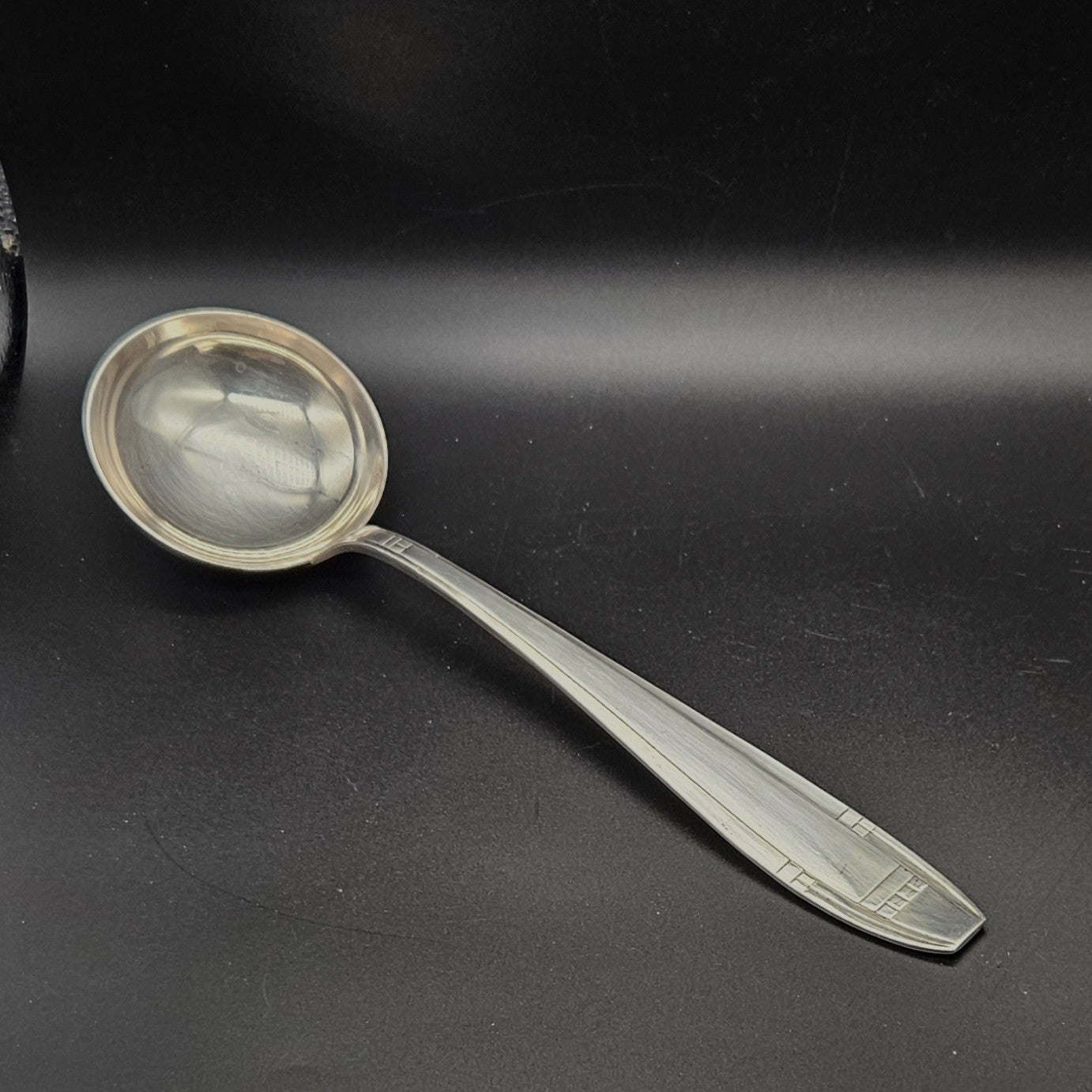 Louche à potage en métal argenté Art Déco, 32,5 cm, vue de face sur fond noir, montrant la cuillère ronde et profonde avec bourrelet périphérique bien marqué et le manche fusiforme effilé à motif géométrique en carrés emboîtés.
