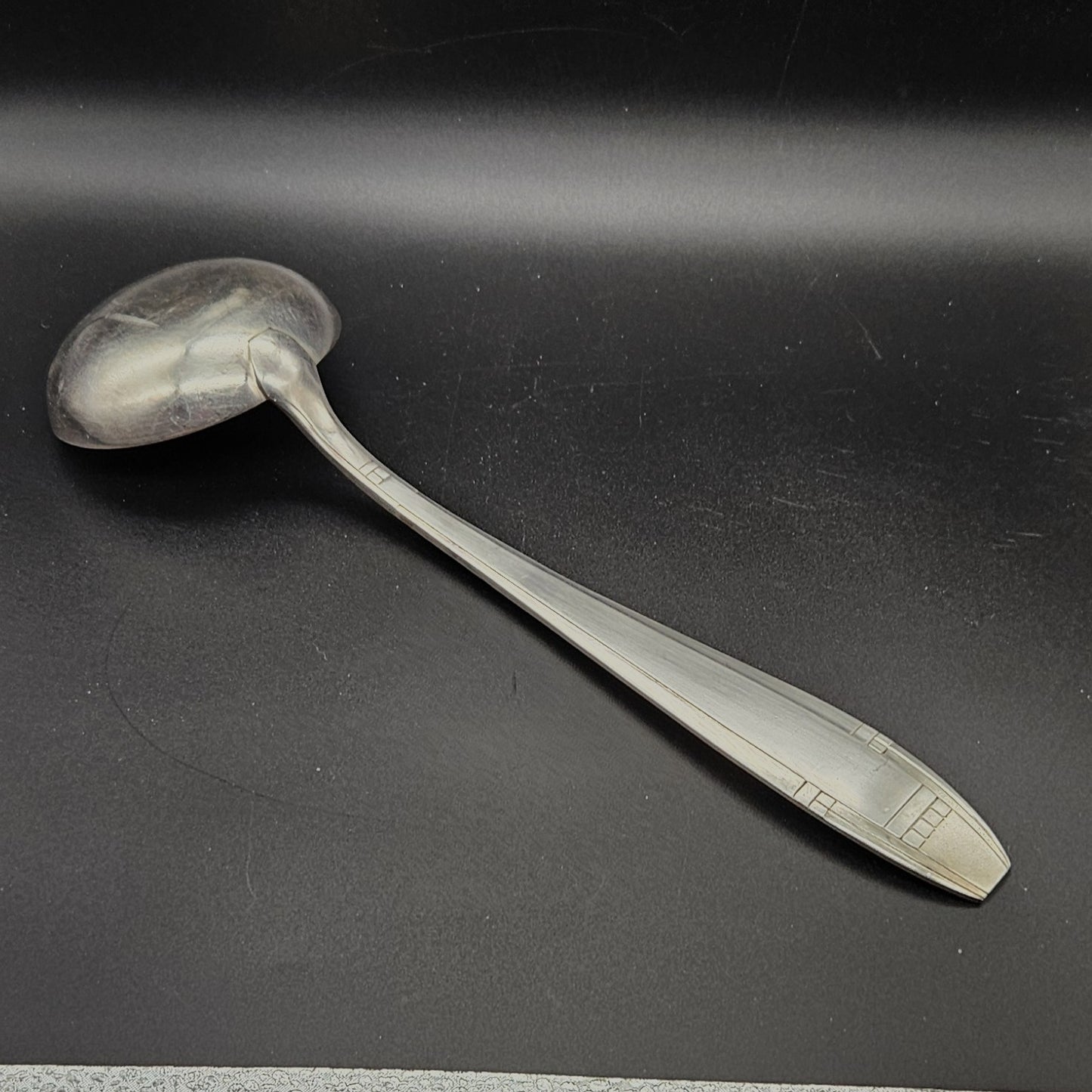 Louche à potage en métal argenté Art Déco, 32,5 cm, vue de dos sur fond noir, montrant le dos de la cuillère ronde et le manche fusiforme effilé à filet central unique avec extrémité à carrés géométriques en relief.