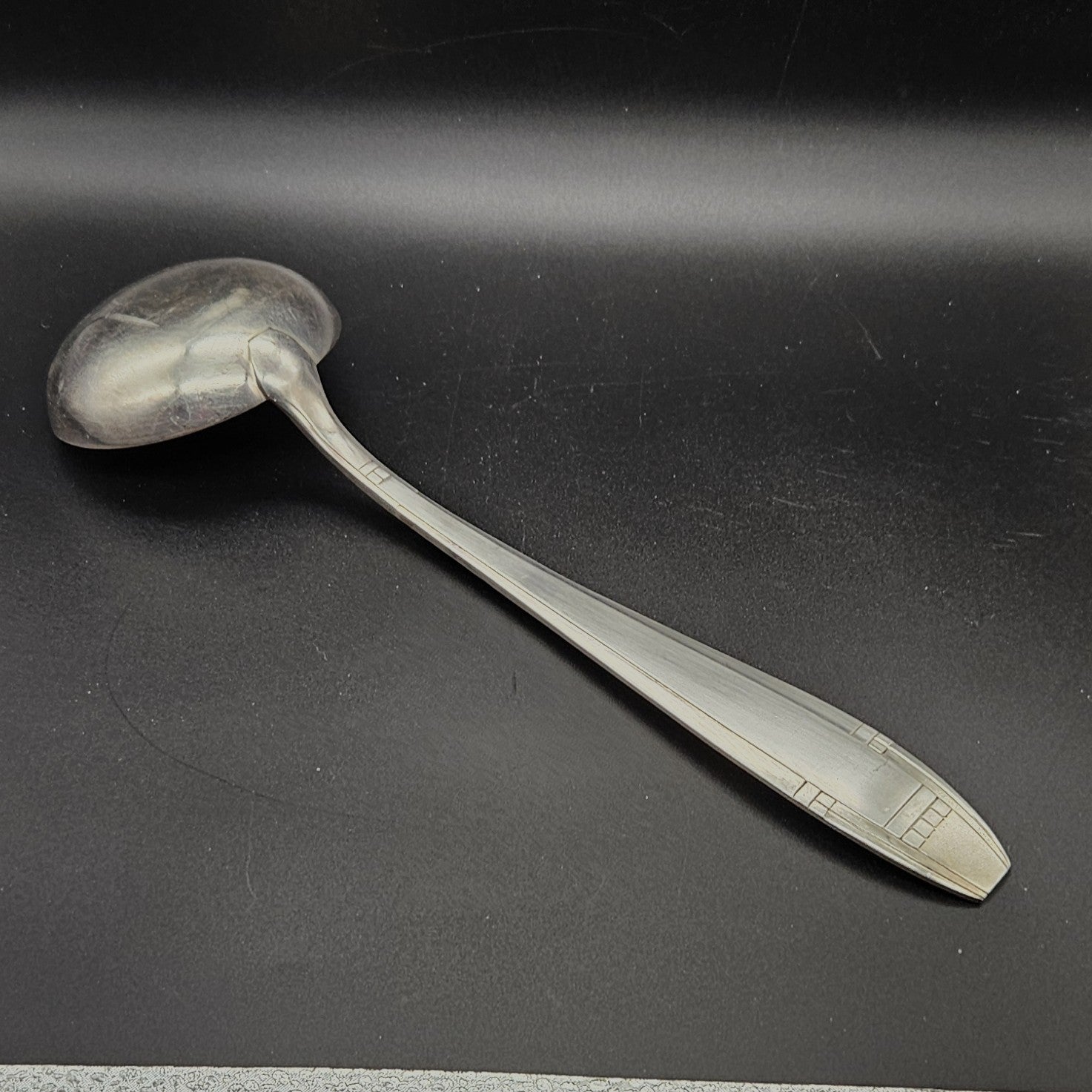 Louche à potage en métal argenté Art Déco, 32,5 cm, vue de dos sur fond noir, montrant le dos de la cuillère ronde et le manche fusiforme effilé à filet central unique avec extrémité à carrés géométriques en relief.