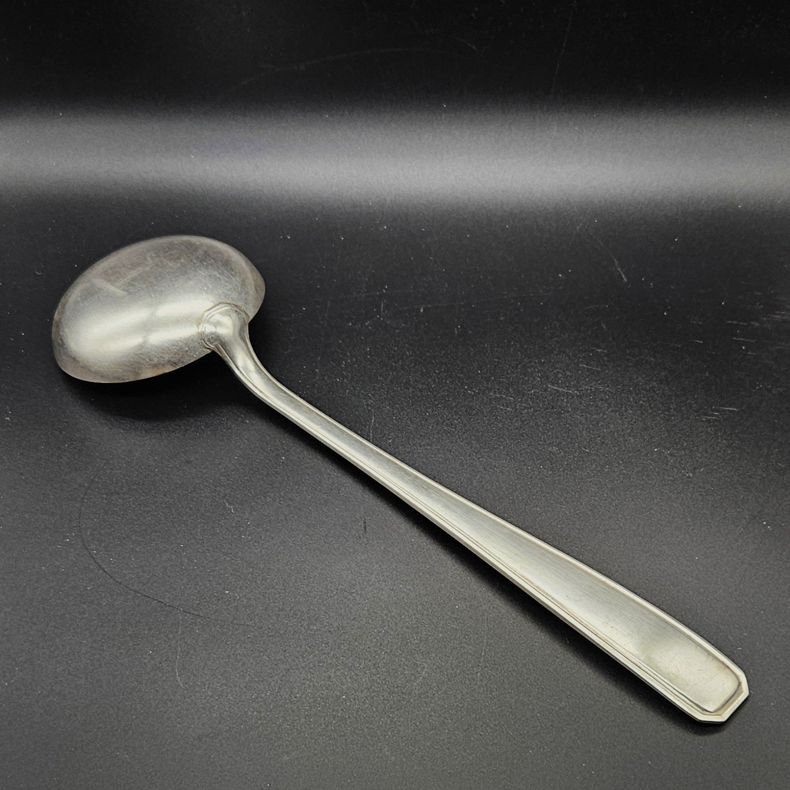 Louche à potage en métal argenté modèle de la Maison Boulenger Paris, 33,7 cm, vue de dos sur fond noir, montrant le dos de la cuillère ronde et le manche rectangulaire à filet périphérique unique avec extrémité à pans coupés octogonaux.