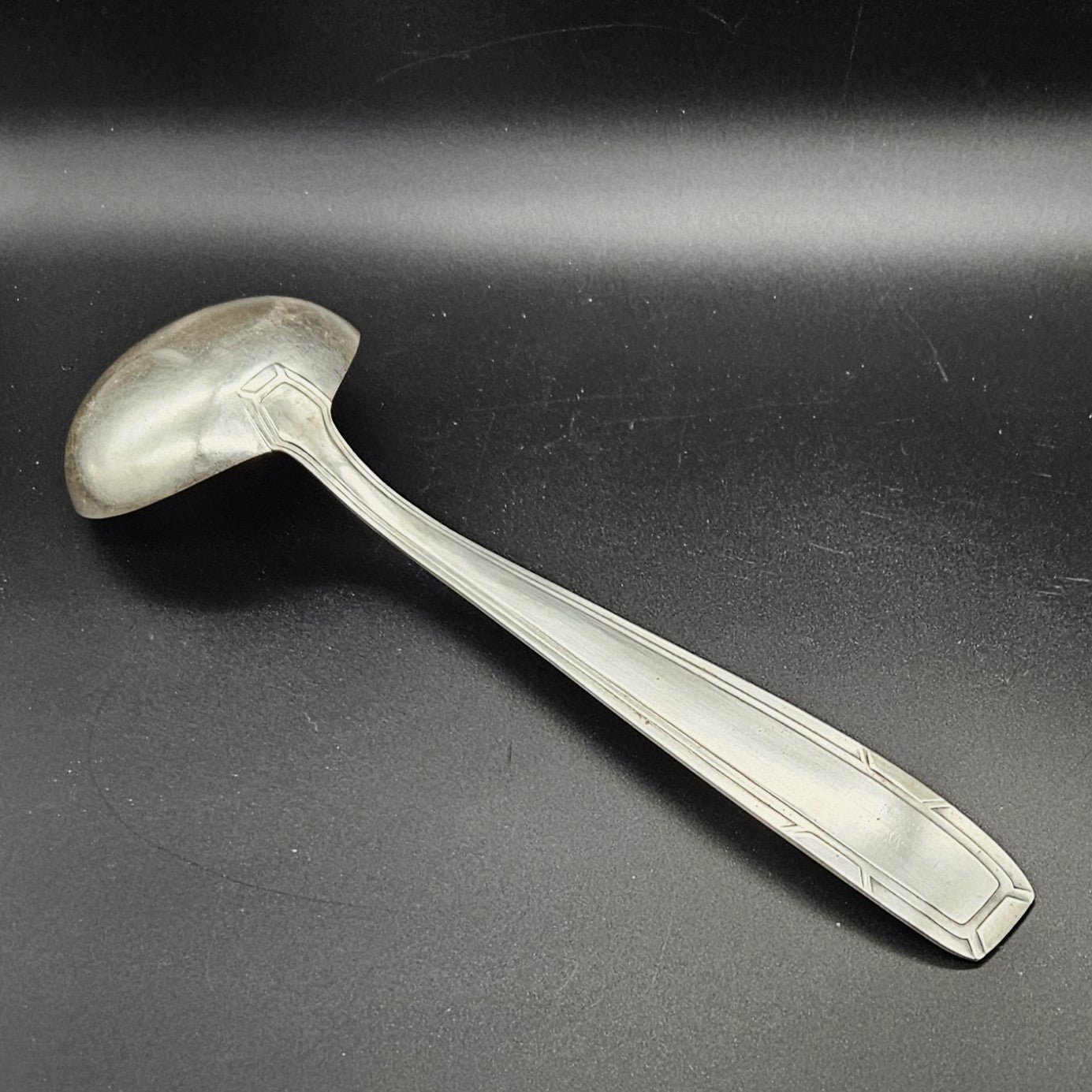 Louche à potage en métal argenté Art Déco, 31 cm, vue de dos sur fond noir, montrant le dos de la cuillère ronde et le manche rectangulaire à double filet latéral convergent avec encoches en V aux angles.