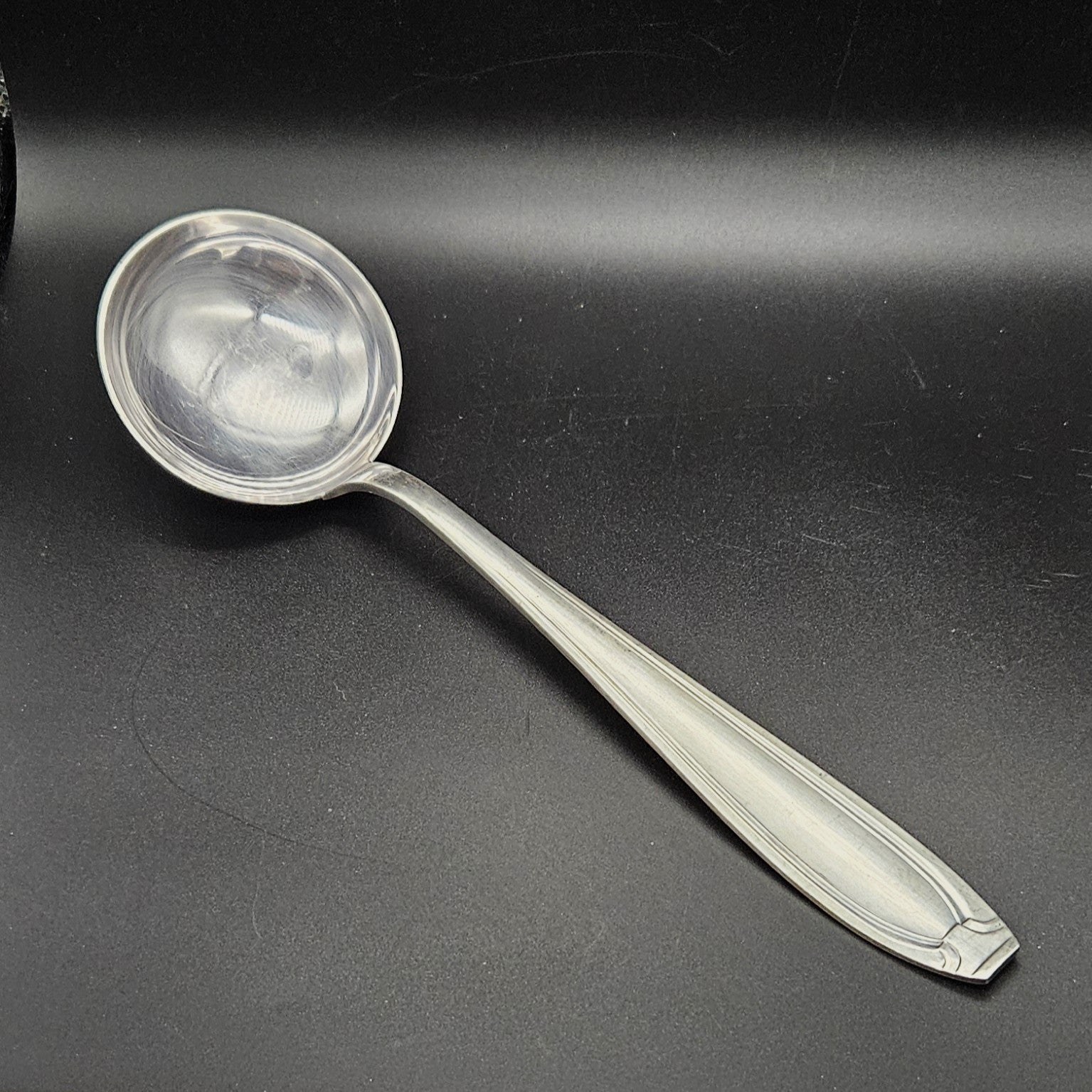 Louche à potage en métal argenté orfèvre Apollo, 32,2 cm, vue de face sur fond noir, montrant la cuillère ronde et profonde avec bourrelet périphérique et le manche fusiforme effilé à double filet latéral convergent.