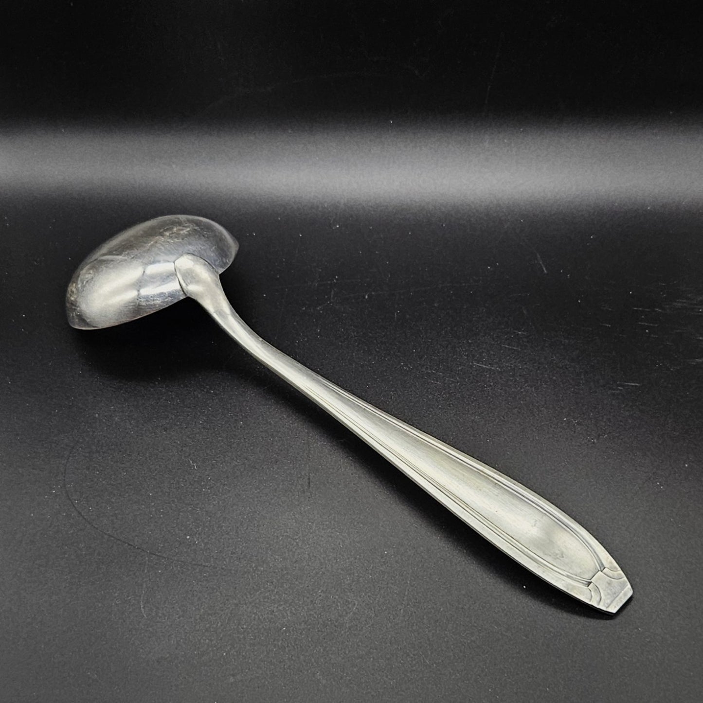 Louche à potage en métal argenté orfèvre Apollo, 32,2 cm, vue de dos sur fond noir, montrant le dos de la cuillère ronde et le manche fusiforme effilé à double filet latéral convergent avec extrémité à pans coupés arrondis.