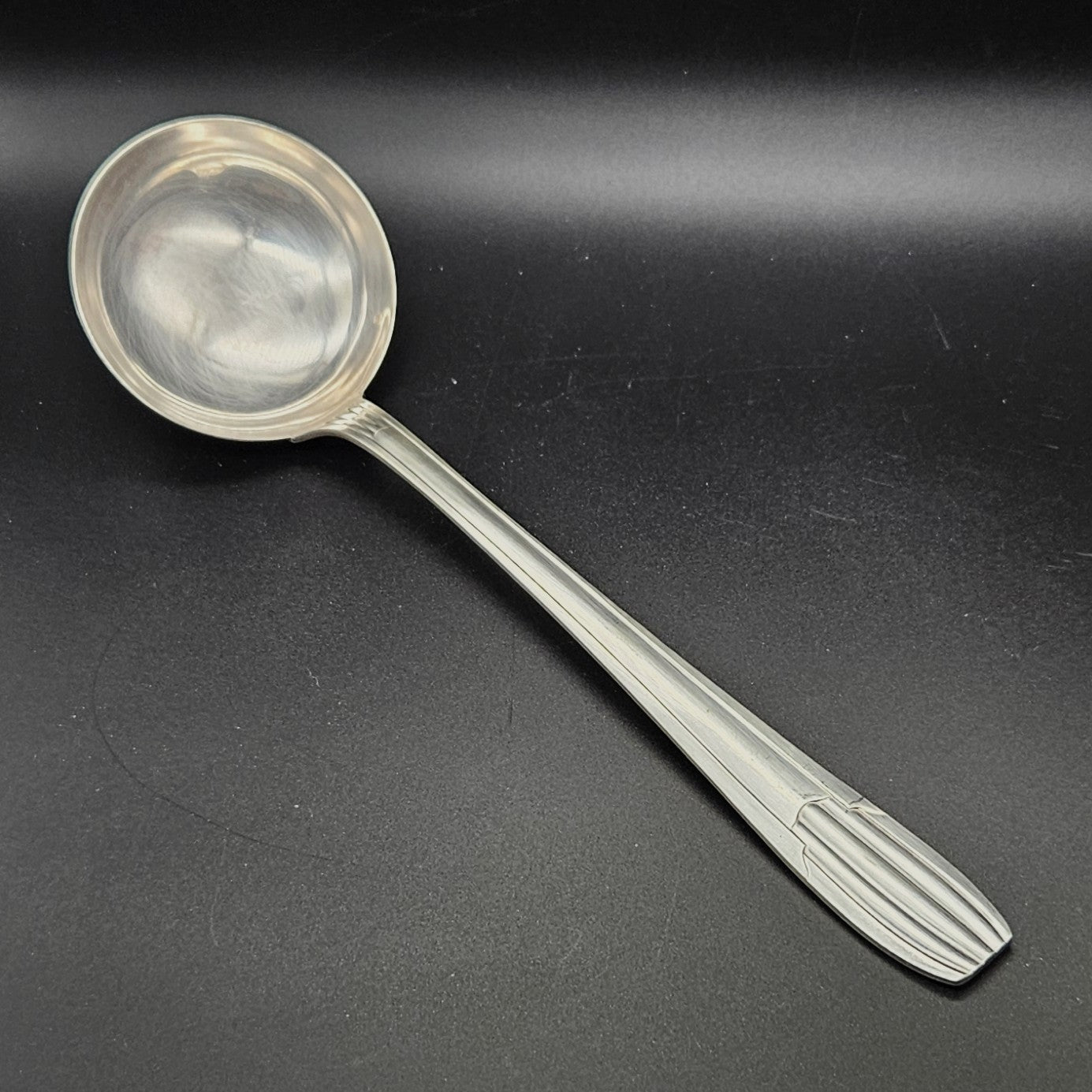Louche à potage en métal argenté Art Déco tardif, 32,3 cm, vue de face sur fond noir, montrant la cuillère ronde et profonde avec bourrelet périphérique et le manche à trois cannelures longitudinales parallèles avec extrémité arrondie à cannelures en éventail.