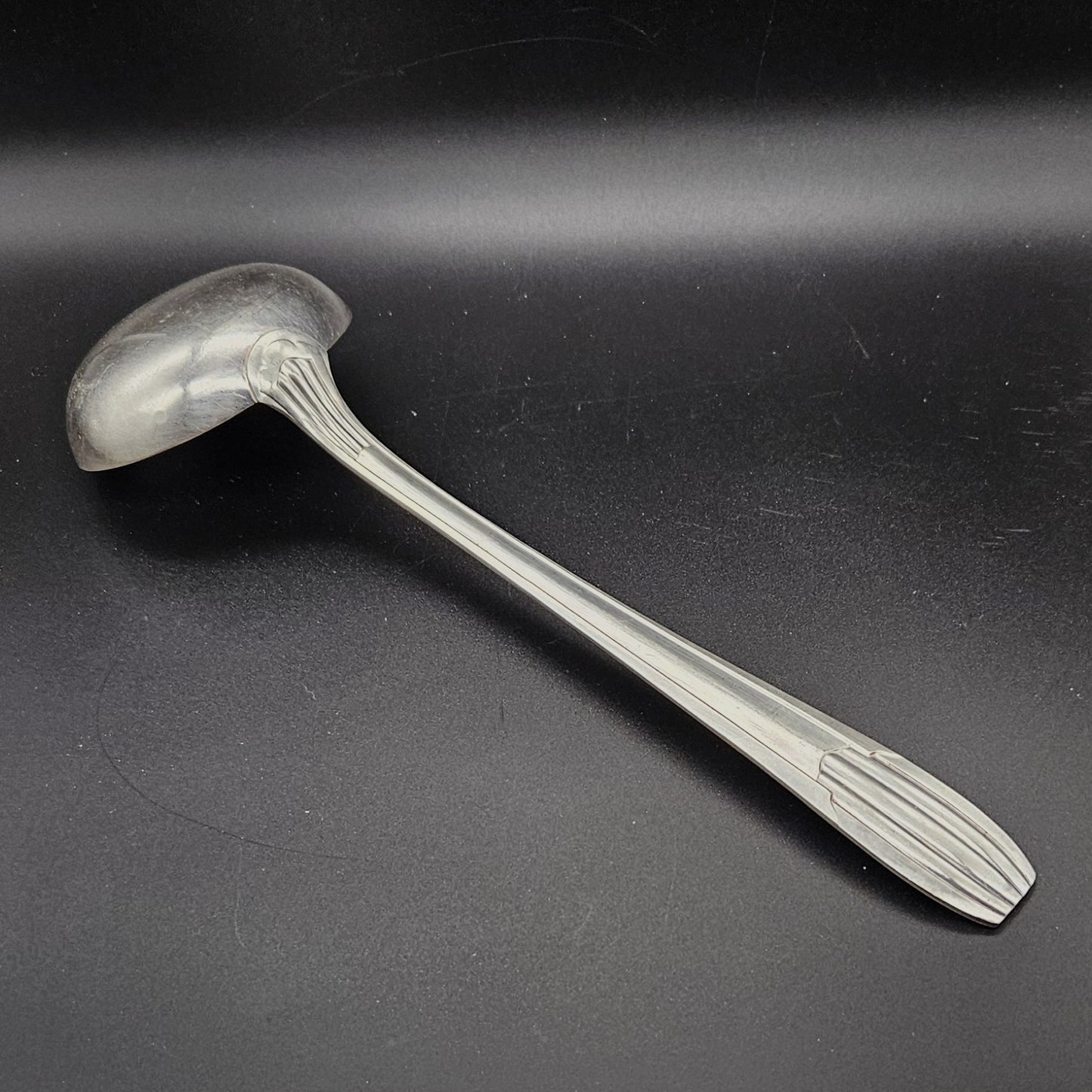 Louche à potage en métal argenté Art Déco tardif, 32,3 cm, vue de dos sur fond noir, montrant le dos de la cuillère ronde et le manche à trois cannelures longitudinales parallèles avec extrémité arrondie à cannelures en éventail.