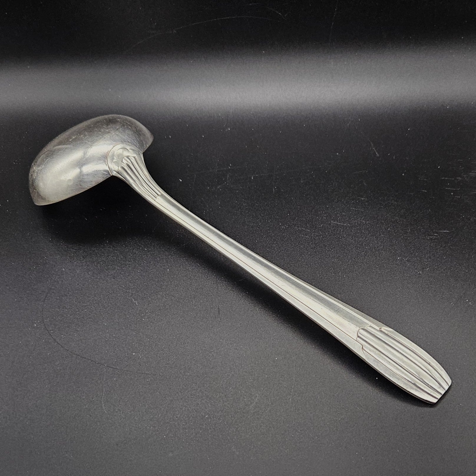 Louche à potage en métal argenté Art Déco tardif, 32,3 cm, vue de dos sur fond noir, montrant le dos de la cuillère ronde et le manche à trois cannelures longitudinales parallèles avec extrémité arrondie à cannelures en éventail.
