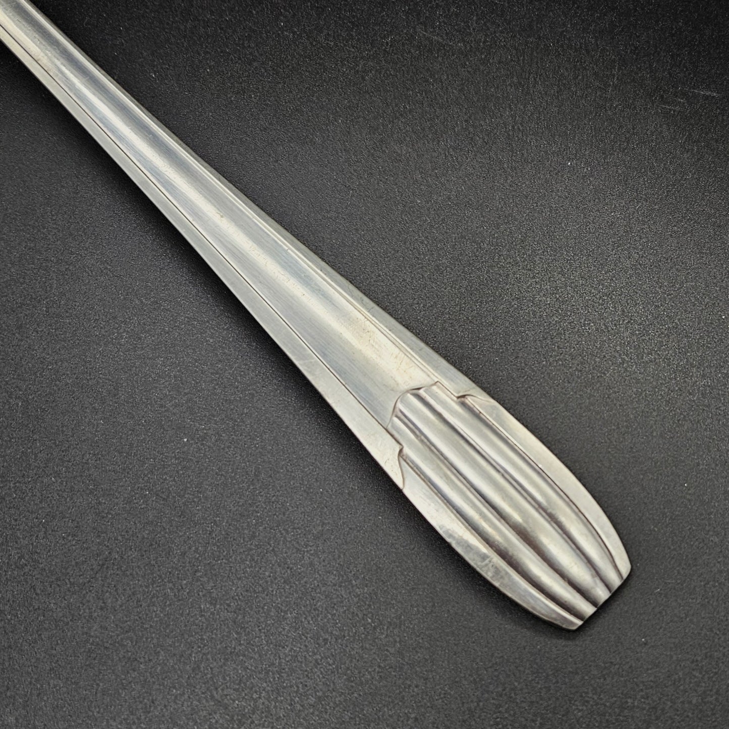 Gros plan sur l'extrémité arrondie du manche de la louche à potage en métal argenté Art Déco tardif, montrant les cinq cannelures en éventail et les trois cannelures longitudinales parallèles du manche, sur fond noir.