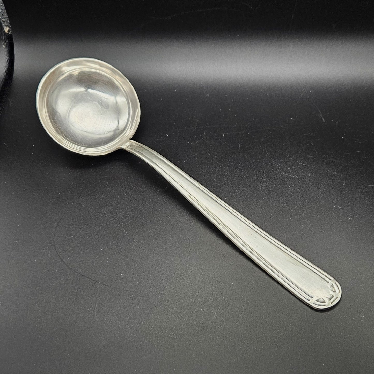 Louche à potage en métal argenté Art Déco, 31,7 cm, vue de face sur fond noir, montrant la cuillère ronde et profonde avec bourrelet périphérique et le manche à double filet latéral parallèle avec extrémité arrondie à motif en arc de cercle à quatre segments.