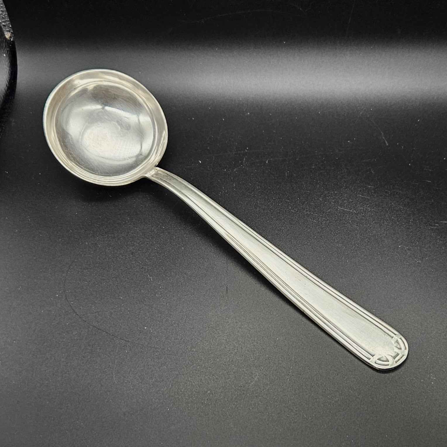Louche à potage en métal argenté Art Déco, 31,7 cm, vue de face sur fond noir, montrant la cuillère ronde et profonde avec bourrelet périphérique et le manche à double filet latéral parallèle avec extrémité arrondie à motif en arc de cercle à quatre segments.