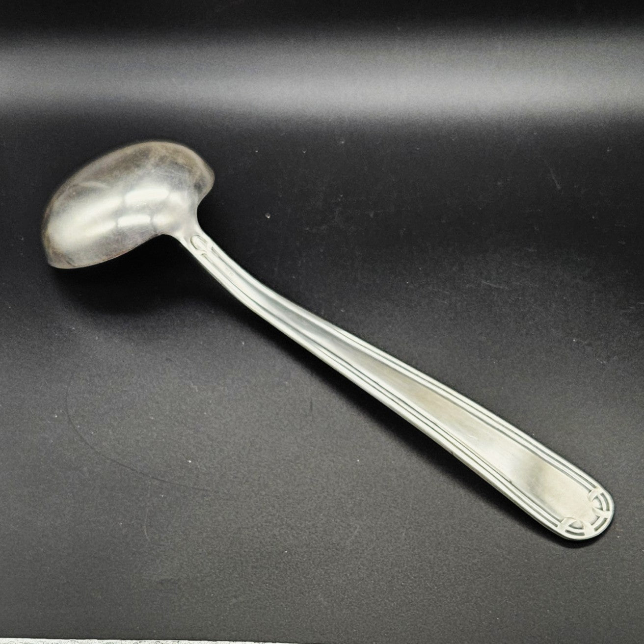 Louche à potage en métal argenté Art Déco, 31,7 cm, vue de dos sur fond noir, montrant le dos de la cuillère ronde avec bourrelet périphérique et le manche à double filet latéral parallèle avec extrémité arrondie à motif en arc de cercle.