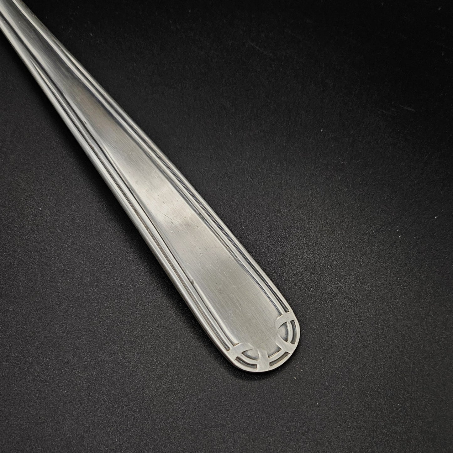 Gros plan sur l'extrémité arrondie du manche de la louche à potage en métal argenté Art Déco, montrant le motif en arc de cercle à quatre segments gravé en creux aux angles et le double filet latéral parallèle, sur fond noir.