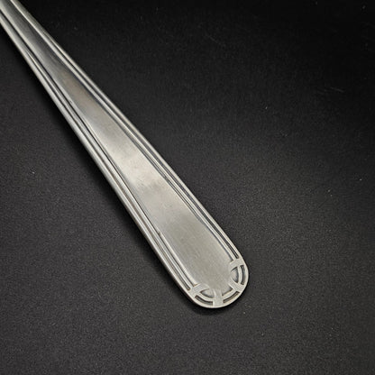 Gros plan sur l'extrémité arrondie du manche de la louche à potage en métal argenté Art Déco, montrant le motif en arc de cercle à quatre segments gravé en creux aux angles et le double filet latéral parallèle, sur fond noir.