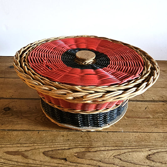 Vue de face de la panière ronde à couvercle en osier tressé tricolore rouge,  noir et naturel, avec bouton de préhension en bois tourné, posée sur une table en bois brut, fond blanc.